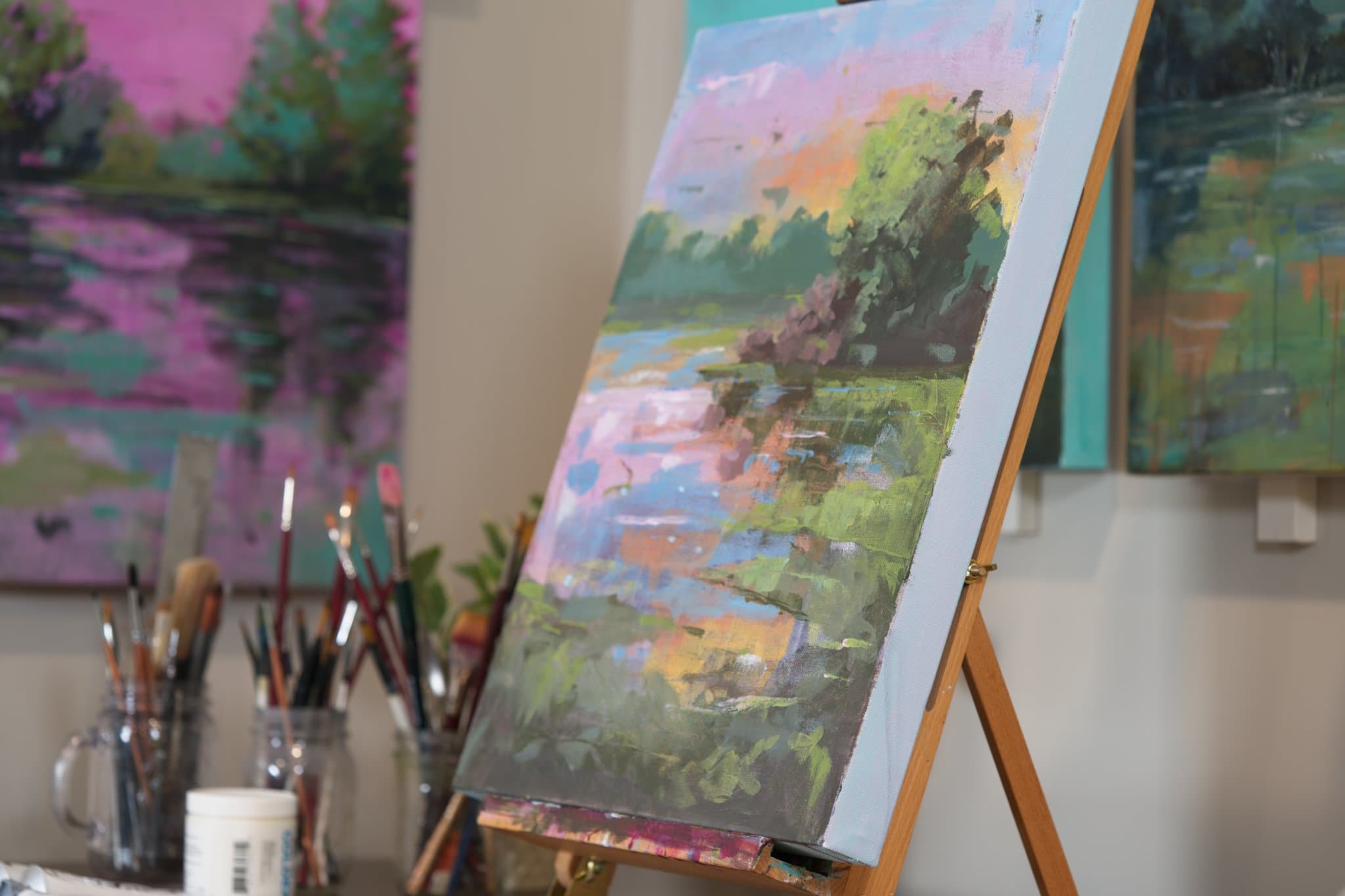 A landscape painting on an easel in Aubrey Cobbett's studio, with brushes in jars beside it
